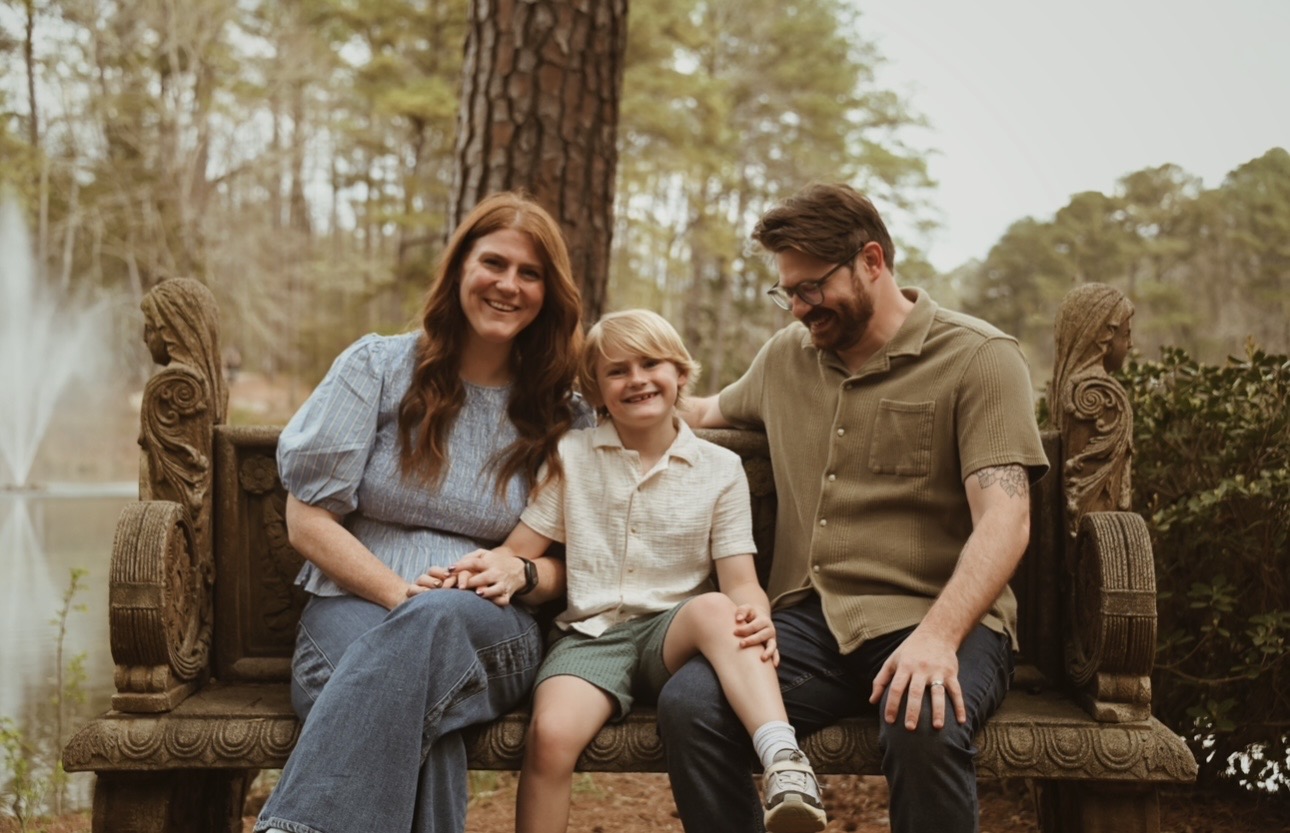Family of three smiling on a carved stone bench by a pond