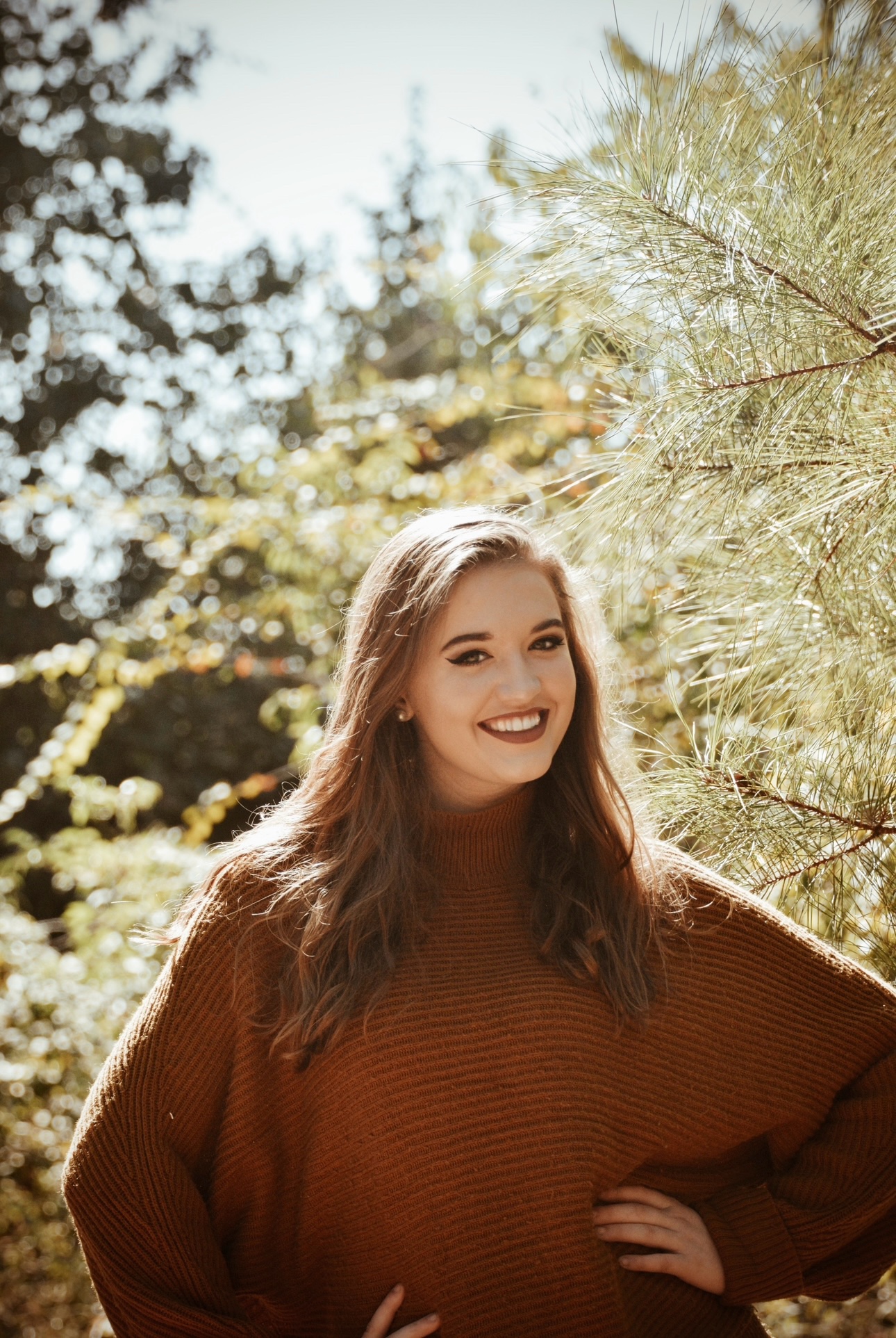 Senior portrait of a young woman in a rust sweater smiling among pines