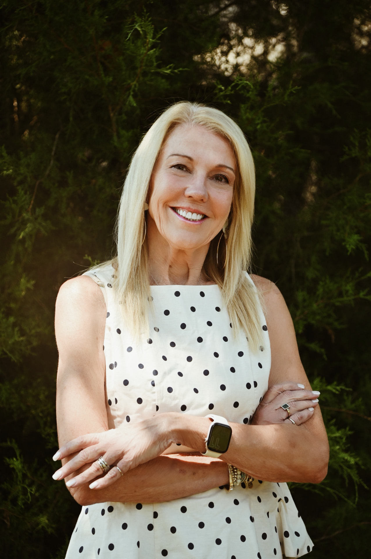 Smiling woman in a cream polka-dot dress with arms crossed in front of evergreen foliage