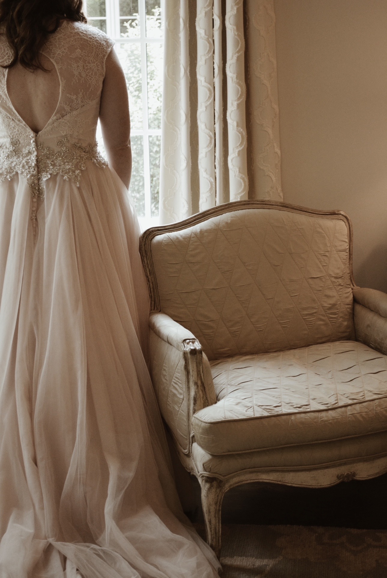 Bride in an elegant gown standing beside a vintage armchair by a window